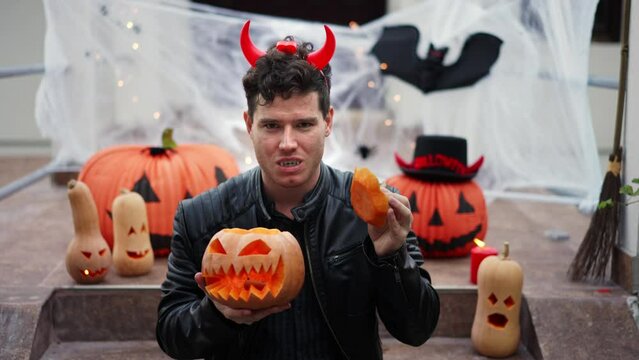 Handsome Young man dressed as a vampire devil with makeup holds and open a pumpkin Jack O Lantern. Halloween props, bats, pumpkins, skulls, spiders, brooms, burning candles, cobwebs. The concept of