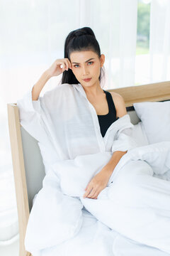 Portrait shot of Millennial Asian sexy female in casual comfort tanktop and shirt sitting under white thick warm blanket posing look at camera smiling after waking up in bedroom in morning at home
