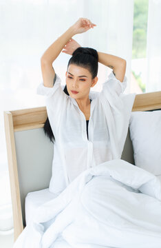 Portrait shot of Millennial Asian sexy female in casual comfort tanktop and shirt sitting under white thick warm blanket posing stretching arm after waking up in bedroom in morning at home.