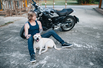 Fair man is sitting on the ground at his motorbike with a dog