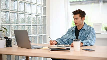 Fototapeta na wymiar Handsome Asian male freelancer remote working in coffee shop, using laptop