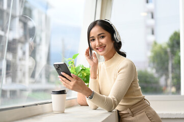 Pretty Asian female is listening to music through headphones, standing by the window.