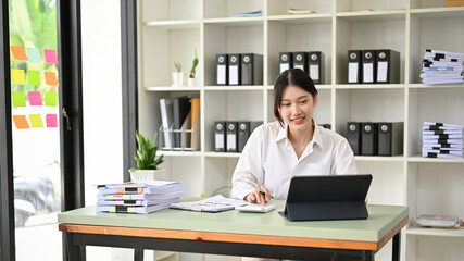 Beautiful Asian female accountant is working art her desk, using calculator and tablet.