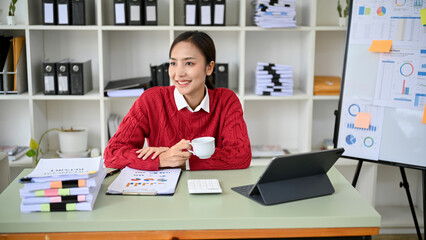 Charming Asian businesswoman having morning coffee at her desk, feeling happy