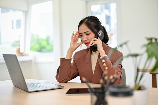 Stressed and thoughtful Asian businesswoman talking on then phone at her desk