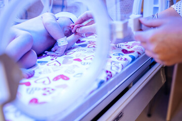 Unrecognizable mother hands caressing and touching her new born baby in intensive care unit in a medical incubator under ultraviolet lamp.