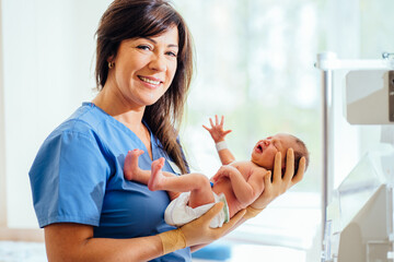 Portrait of maternity nurse or lactation consultant wearing blue scrubs holding newborn baby girl in diaper in hospital room.