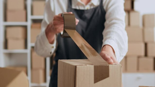 Packing a box A parcel warehouse worker packs a box with brown tape, Packing a brown box with sticky tape, a small business worker works in a warehouse e-commerce online store.
