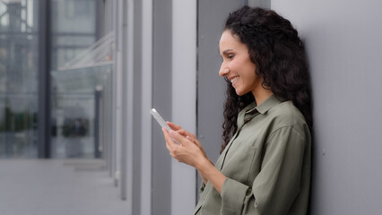 Side view Caucasian Hispanic woman 30s businesswoman holding cell phone standing near wall building outdoors success good news mobile app win surprised lady celebrating victory on smartphone triumph