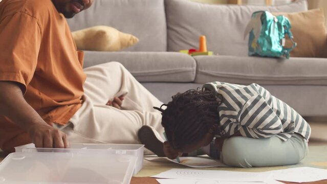 Black man spending time with 3 year old daughter, sitting on rug in living room and drawing rainbow with colorful crayons