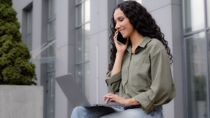 Side view multitasking businesswoman freelancer worker lady sitting outdoor outside talking mobile phone typing laptop answer call ordering booking ecommerce shopping online with wireless computer app