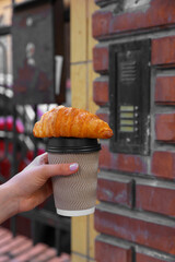 Woman holding croissant and paper cup in hand outdoors, closeup