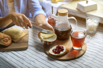 Woman spreading butter on bread near glass mug of aromatic tea indoors, closeup