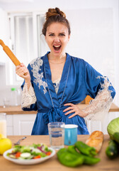 Angry woman screaming and waving rolling pin in home kitchen