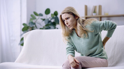 Fototapeta premium blonde woman having pain in back while sitting on couch in modern living room