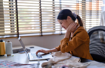 Young professional clothes fashion designer sitting near sewing machine use laptop computer and tablet pc to reference and she feel fatigue due to work late and sleepy on work bench