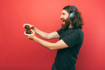 Side view photo of amazed bearded man playing games with joystick and screaming over pink...