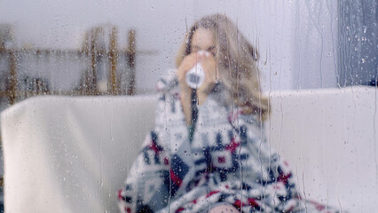 sick woman sitting under colorful blanket and drinking tea behind wet window with rain drops