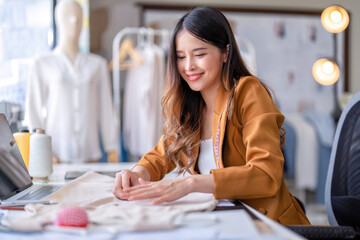 Young professional clothes fashion designer sitting near sewing machine use laptop computer and tablet pc to reference and concentrate on her work pattern