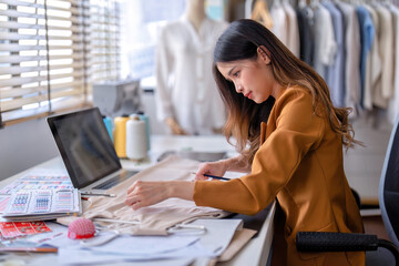 Young professional clothes fashion designer sitting near sewing machine use laptop computer and tablet pc to reference and draw the design on the paper pattern