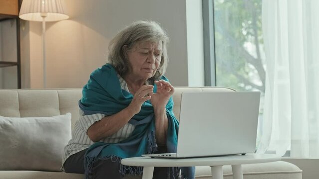 Elderly woman sitting on sofa at home and speaking via online video call on laptop