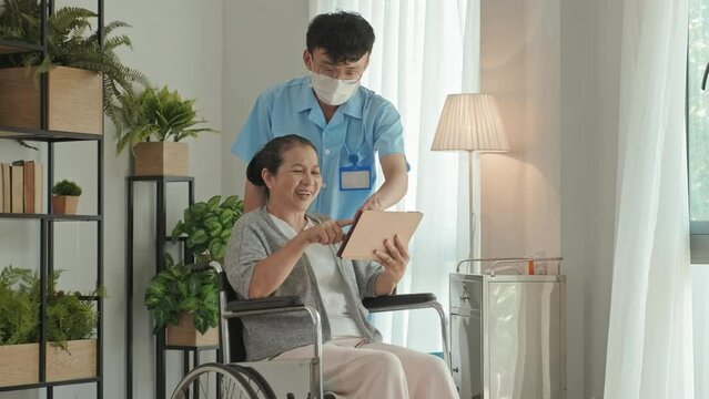 Medium shot of senior Asian woman on wheelchair smiling and using digital tablet with assistance of male nurse in scrubs and mask while spending time in care home