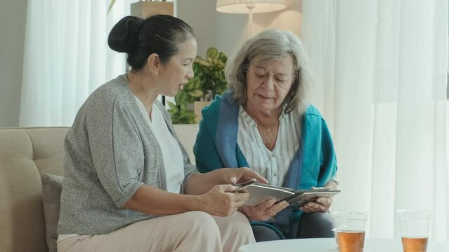 Medium shot of two senior women sitting on sofa in living room, watching photos in album and talking