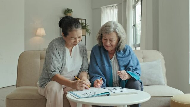 Medium tracking shot of cheerful Asian and Caucasian elderly women sitting on sofa in nursing home, chatting and laughing while solving crossword together