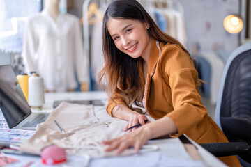 Young professional clothes fashion designer sitting near sewing machine use laptop computer and tablet pc to reference design smiling portrait to camera