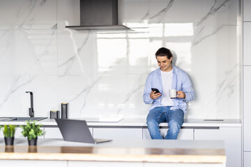 Young man using smartphone, cooking salad in modern kitchen, searching in internet, looking at phone screen