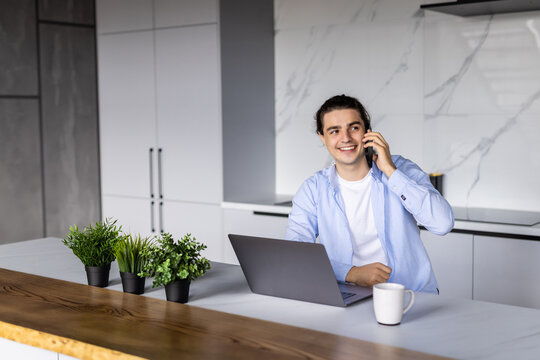 Gig economy concept. Young positive man freelancer working remotely at home, looking at laptop and consulting client via cellphone, sitting at kitchen