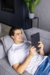 Young man with beard lying on the sofa and holding tablet in hands. Man indoor with laptop tablet. Hipster man relaxing and looking to the tablet. Young man with tablet.