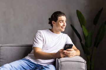Portrait of an attractive smiling young man wearing casual clothes sitting on a couch at the living room, using mobile phone