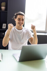 Excited young man winner sit at home table looking at laptop screen celebrating online success...