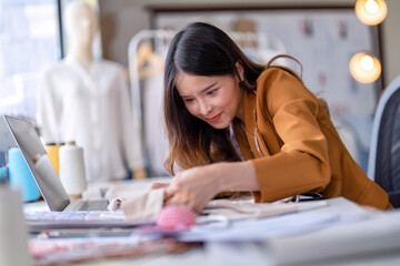 Young professional clothes fashion designer sitting near sewing machine use laptop computer and tablet pc to reference and concentrate on her work pattern