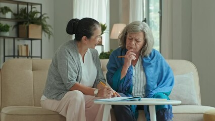 Medium shot of Asian and Caucasian elderly women sitting on couch, speaking and solving crossword together while spending free time in nursing home