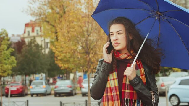 Attractive young woman leather jacket with checkered scarf waiting under rain with an blue umbrella talking on phone in windy weather. Cars and people on blurred background. Concept of city life