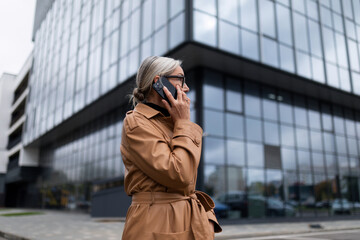Fototapeta premium Business senior adult woman in an autumn coat on the background of the glass facade of the office center speaks on a mobile phone, 50 year old European woman, business strategy concept