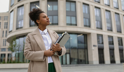 successful ambitious woman of African American appearance in business clothes with a laptop in her hands against the backdrop of a city building, business strategy concept