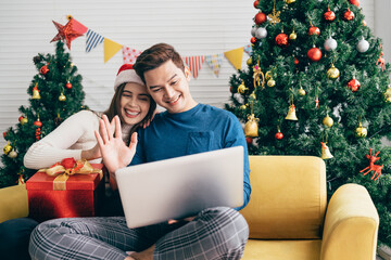 Young happy Asian couple sitting together video call on a laptop with friends or parents thanks for a gift received during the Christmas holiday with a decorated Christmas tree in the background.