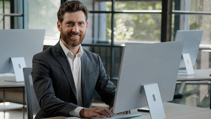Portrait adult bearded freelancer worker man at office working typing laptop smiling at camera. Portrait satisfied 40s businessman browsing laptop at workplace happy with new job in modern company