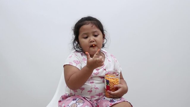 Adorable Asian little girl sitting while eating snack. Isolated on white background
