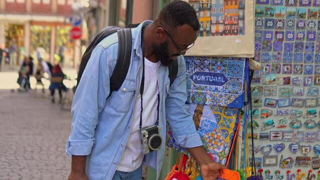  Young african american traveler man with a camera choosing the souvenirs at the typical souvenir shop. 