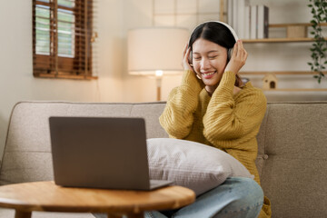 Quiet Asian young woman in modern wireless headphones sits comfortably on the sofa, listening to music with calm happiness.