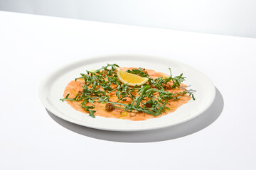 Salmon carpaccio with arugula on white background in summer day. Fish appetizer - salmon carpaccio in fine dining with shadows. Elegant carpaccio of raw salmon and sauce. Italian food. Aesthetic menu.