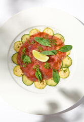 Beef carpaccio with zucchini and cucumber on white table with sunlight shadows. Appetizer of meat - beef carpaccio on summer dining. Sliced raw meat in italian style. Aesthetic menu.