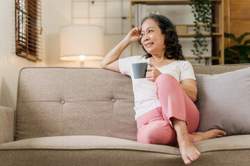 Old pretty asian woman sitting on the sofa with coffee looking out of windows.