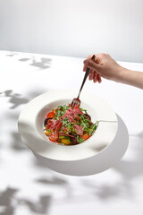 Meat salad and female hand. Woman eating elegant summer salad. Composition with steak salad and fork holding person. Trendy summer food on white background with female hand.