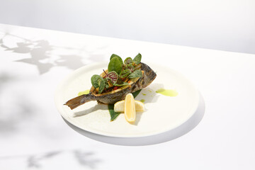 Whole roasted fish with spinach leaves on white plate. Grilled dorado on light background in summer menu. Healthy food - Roasted dorado fish. Concept menu with hard shadows.