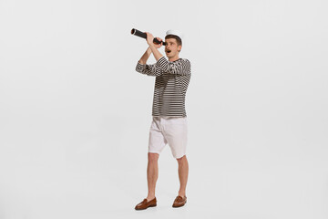 Portrait of young man, sailor in striped shirt looking in spyglass isolated over white background. Curious person - obrazy, fototapety, plakaty